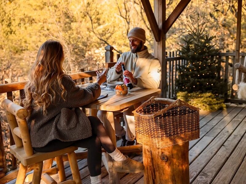 Couple assis à une table en bois sur une terrasse en forêt, partageant un moment convivial.