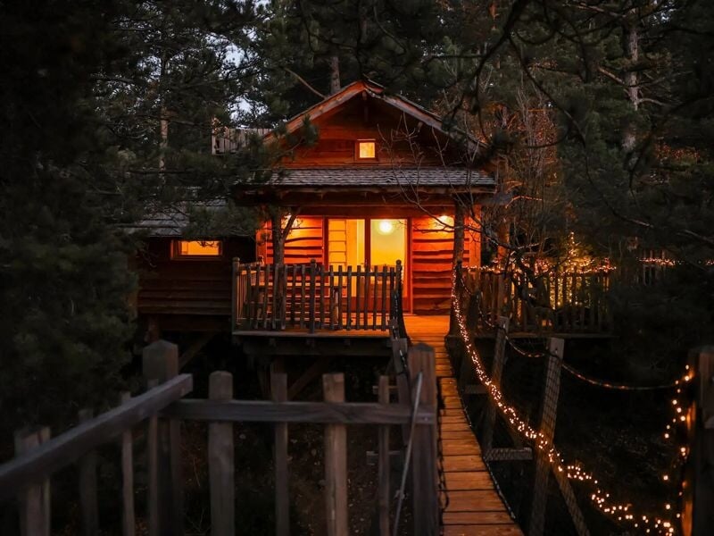 Cabane en bois éclairée la nuit avec un pont suspendu décoré de guirlandes lumineuses.
