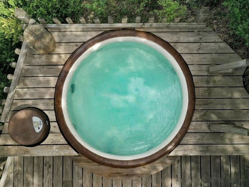 Jacuzzi circulaire en bois sur une terrasse en bois, entouré de végétation verte.
