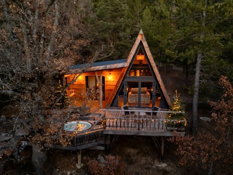 Cabane en bois en forme de A avec terrasse, jacuzzi et guirlandes lumineuses dans la forêt.