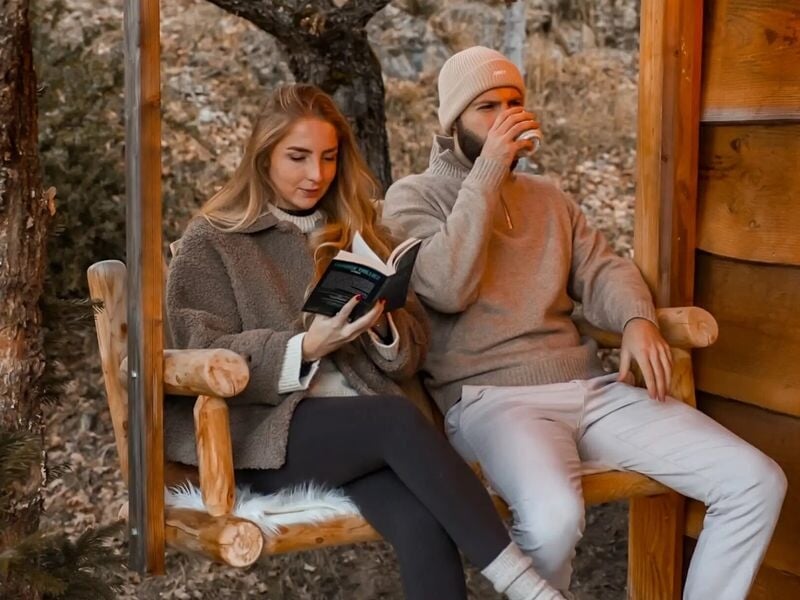 Couple assis sur une balançoire en bois, l'homme boit un verre pendant que la femme lit un livre.