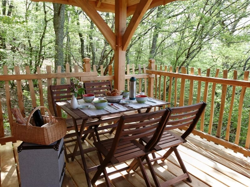 Terrasse en bois avec table et chaises, petit déjeuner posé, entourée d'arbres verts et ombragés.