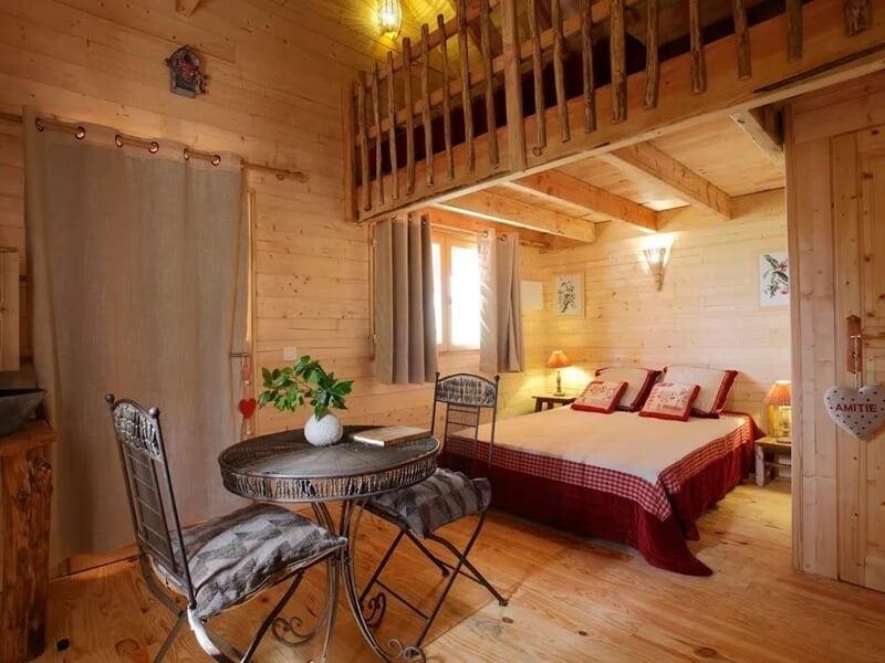 Chambre en bois avec lit double rouge, table ronde et chaises, ambiance chaleureuse et rustique.