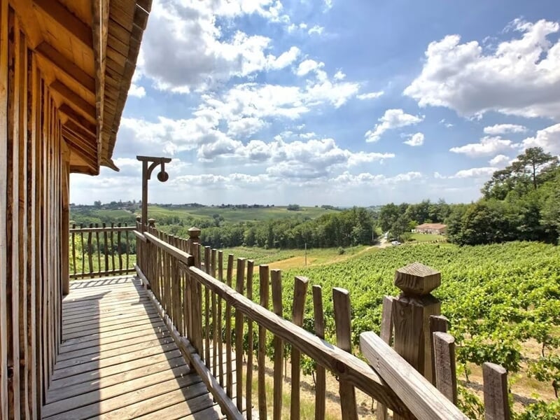 Balcon en bois avec vue sur un paysage vallonné de vignes sous un ciel bleu parsemé de nuages.