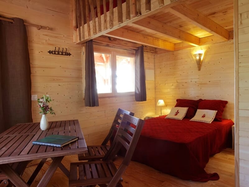 Chambre en bois avec lit rouge, table en bois et chaises, lumière naturelle entrant par la fenêtre.