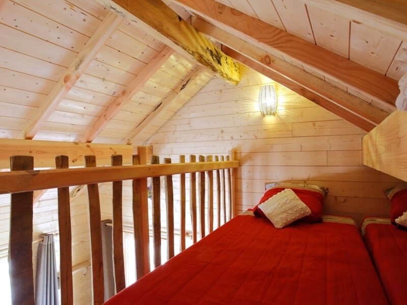 Mezzanine en bois avec lit simple rouge, rambarde en bois, lumière douce et ambiance cosy.