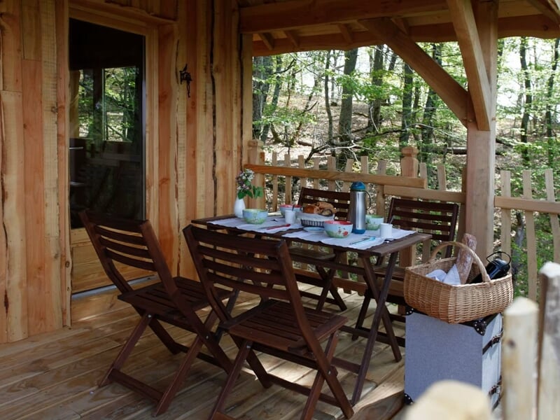 Terrasse en bois avec table et chaises, panier de pique-nique posé, entourée d'arbres verts.