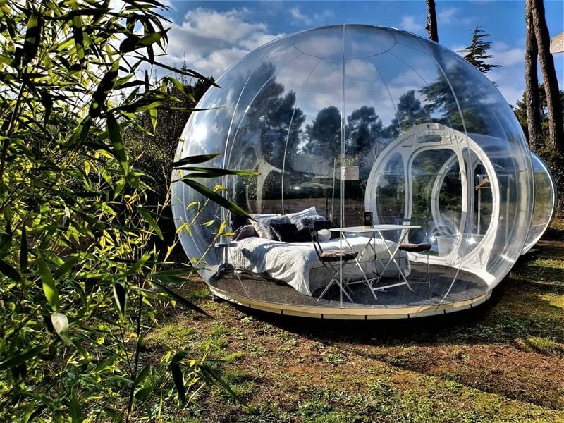 Bulle transparente installée en pleine nature avec lit et table, entourée d'arbres sous un ciel partiellement nuageux.