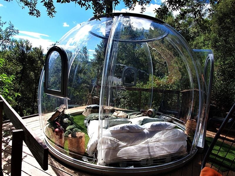Bulle transparente avec lit double à l'intérieur, posée sur une terrasse en bois entourée de végétation dense.