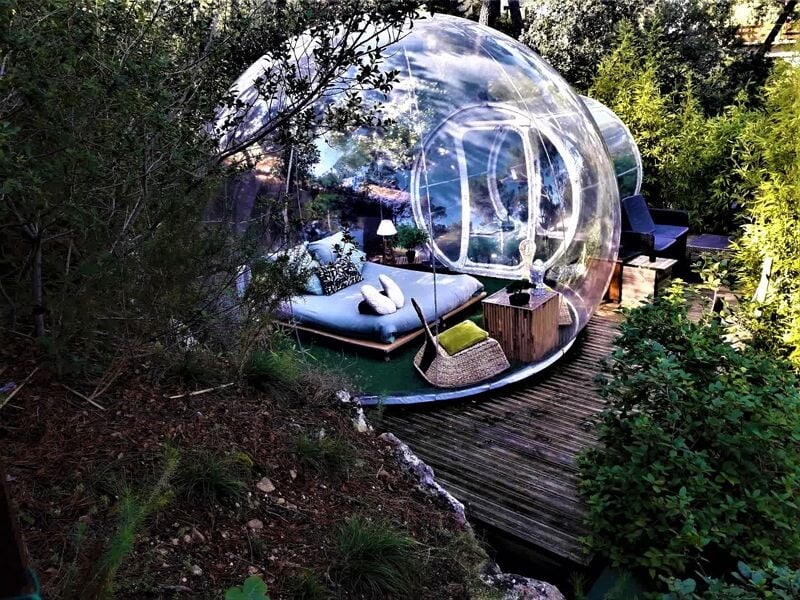 Bulle transparente avec espace extérieur aménagé, canapé et coussins, entourée d'arbres et de buissons verts.