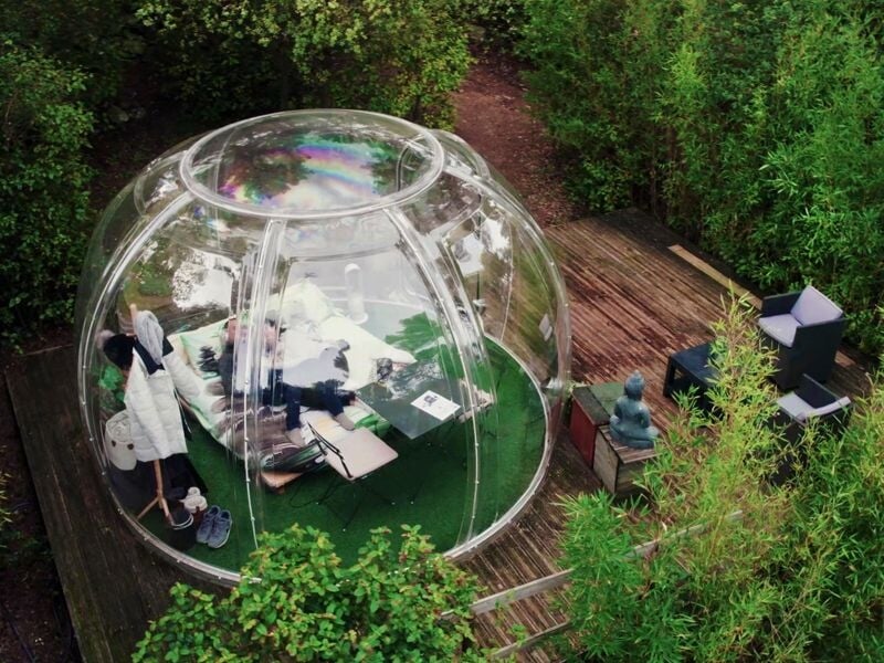 Bulle transparente vue de haut avec lit à l'intérieur, située sur une terrasse en bois au milieu d'une forêt dense.