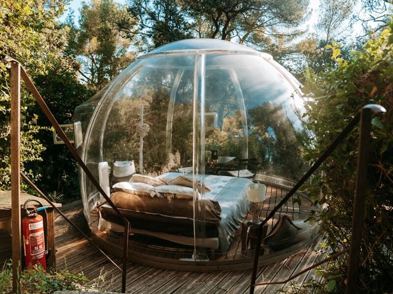 Bulle transparente avec lit double à l'intérieur, posée sur une terrasse en bois, entourée d'arbres et d'un extincteur rouge.