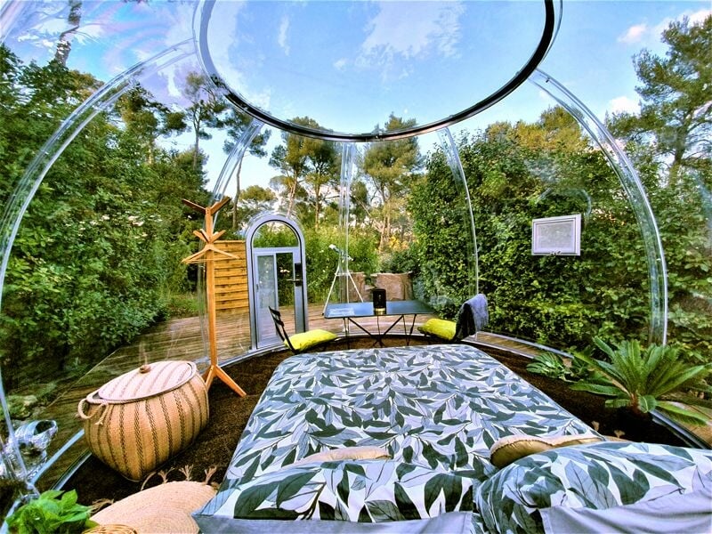 Intérieur d'une bulle transparente avec lit double, table et chaises, entourée de végétation luxuriante sous un ciel bleu.