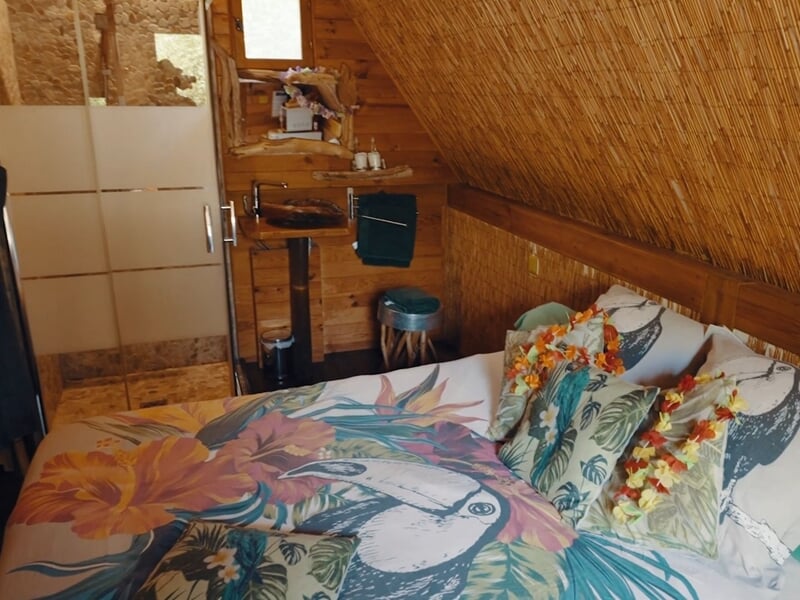 Intérieur d'une chambre en bois avec lit décoré de coussins à motifs tropicaux et guirlandes de fleurs.