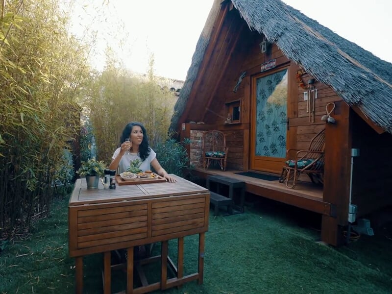 Femme assise à une table en bois dans un jardin entouré de bambous devant une petite cabane.