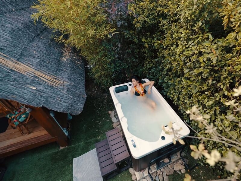 Femme dans un jacuzzi extérieur entouré de végétation dense et d'une cabane en bois à côté.