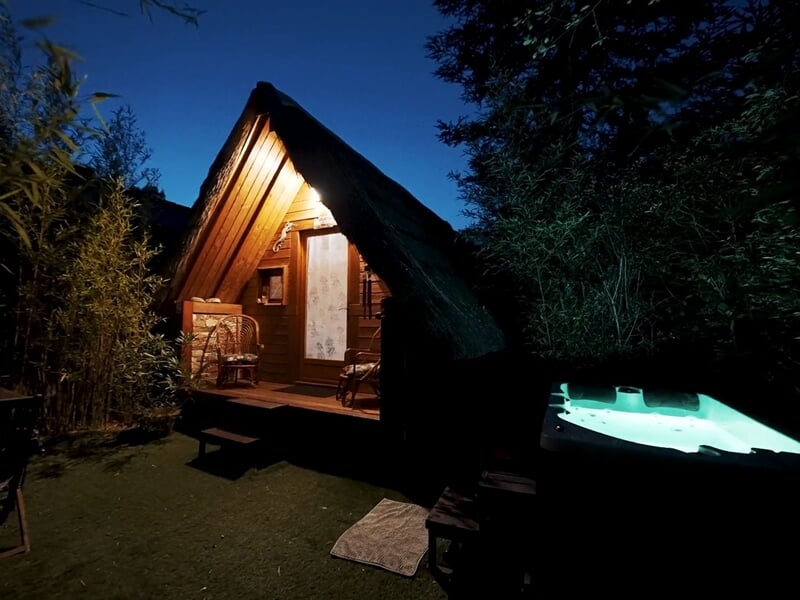 Cabane en bois éclairée la nuit avec jacuzzi extérieur illuminé dans un jardin verdoyant.