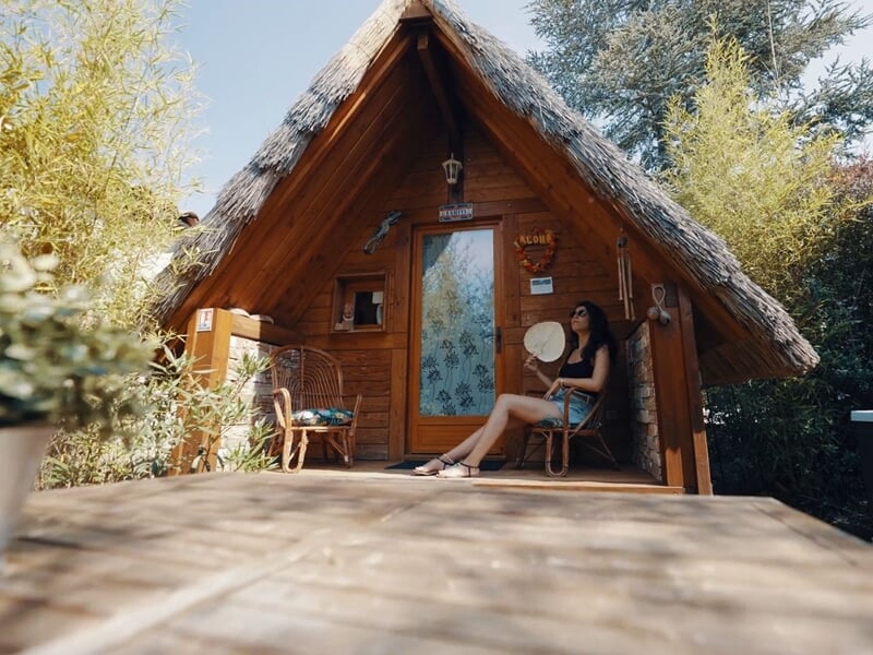 Femme assise sur une chaise en bois sur la terrasse d'une cabane en bois entourée de verdure.