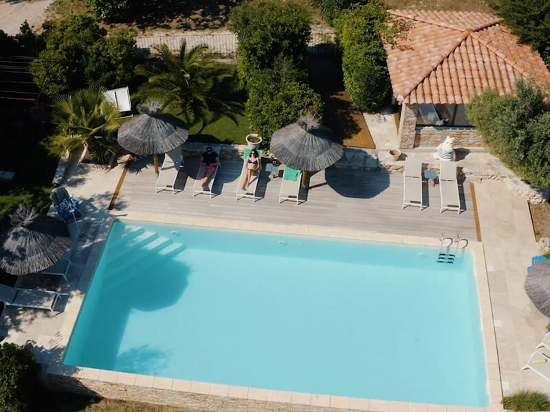 Piscine extérieure entourée de transats et parasols en paille sous un ciel bleu et arbres verts.