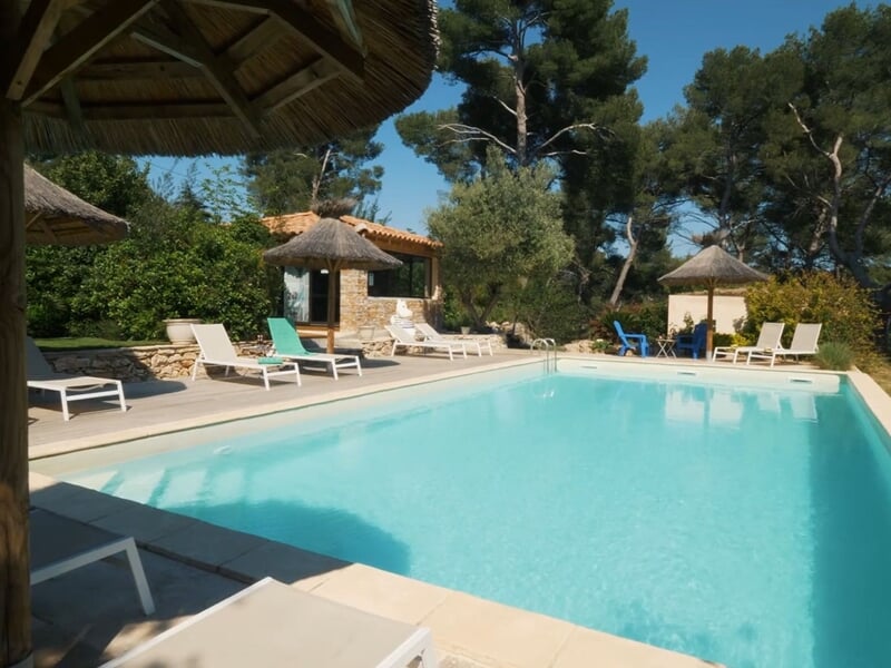 Piscine claire avec transats blancs et parasols en paille dans un jardin arboré ensoleillé.