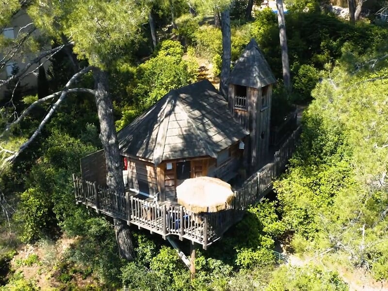 Cabane en bois perchée dans les arbres avec terrasse et parasol, entourée de végétation dense et verdoyante.