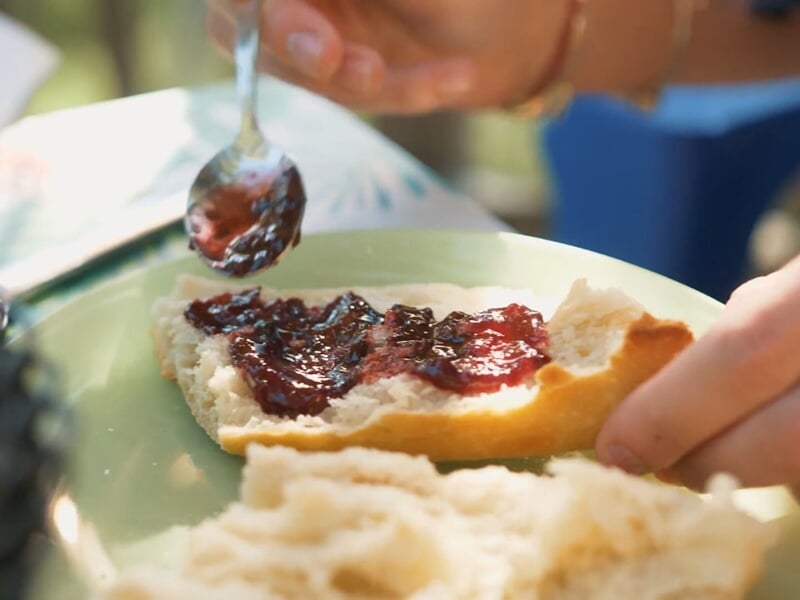 Main étalant de la confiture rouge sur une tranche de pain blanc sur une assiette verte.