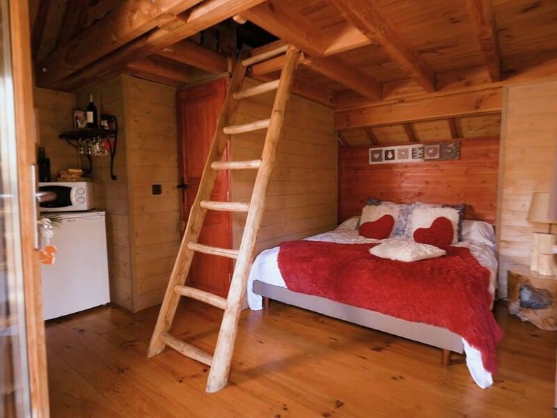 Intérieur chaleureux d'une cabane en bois avec lit double, couverture rouge et échelle en bois.