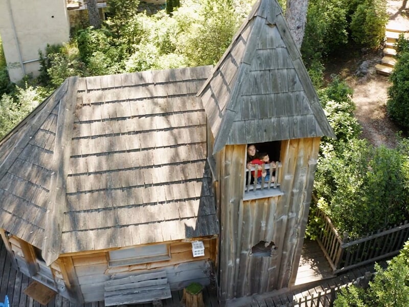 Cabane en bois vue de haut avec toit en ardoise et petite tour, deux enfants regardent par la fenêtre.