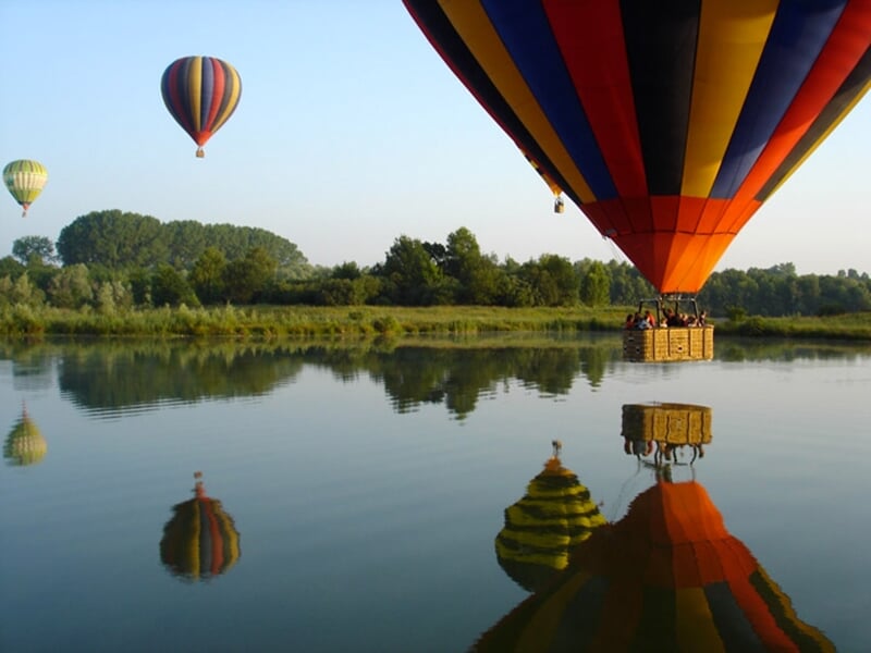 Montgolfière colorée survolant un lac calme, avec des reflets parfaits sur l'eau et d'autres montgolfières en arrière-plan.