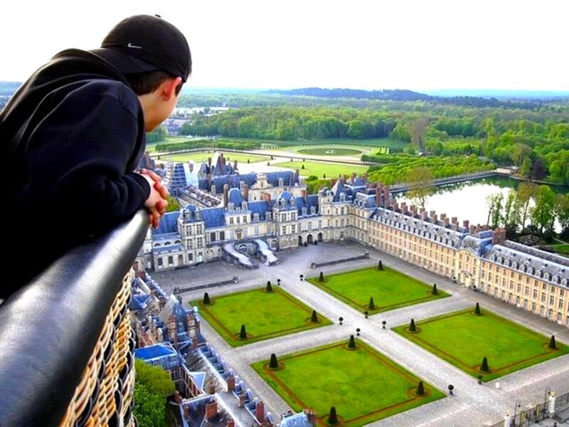 Un homme regardant depuis la nacelle d'une montgolfière une vue plongeante sur un château majestueux avec ses jardins symétriques et ses allées verdoyantes.