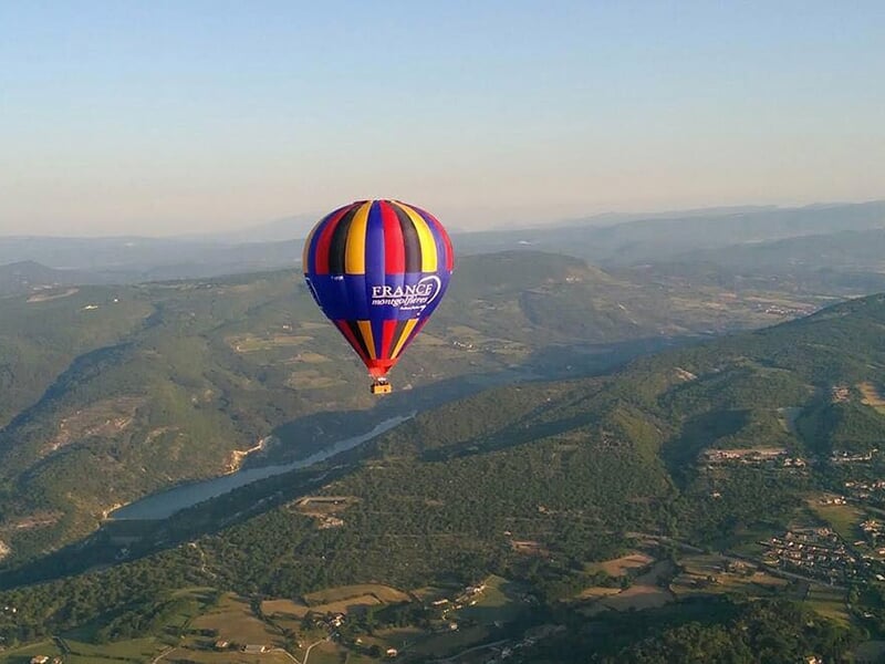Montgolfière colorée survolant une vallée avec une rivière sinueuse et des collines verdoyantes au lever du soleil.