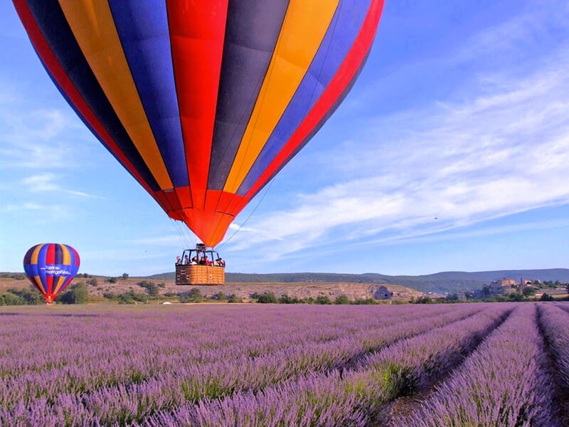 Montgolfière colorée en vol rasant un champ de lavande en fleurs avec une autre montgolfière visible au loin sous un ciel bleu.