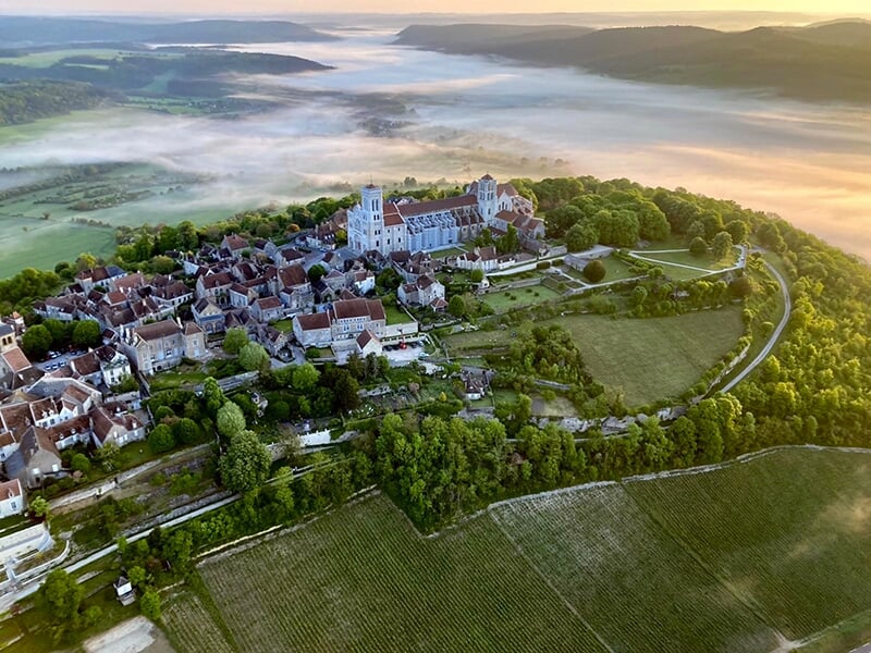 Vue aérienne d'un village médiéval entouré de verdure et de brume au lever du soleil.