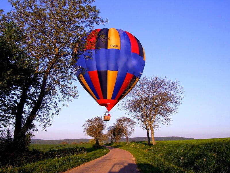 Montgolfière colorée flottant au-dessus d'un chemin bordé d'arbres sous un ciel bleu clair.
