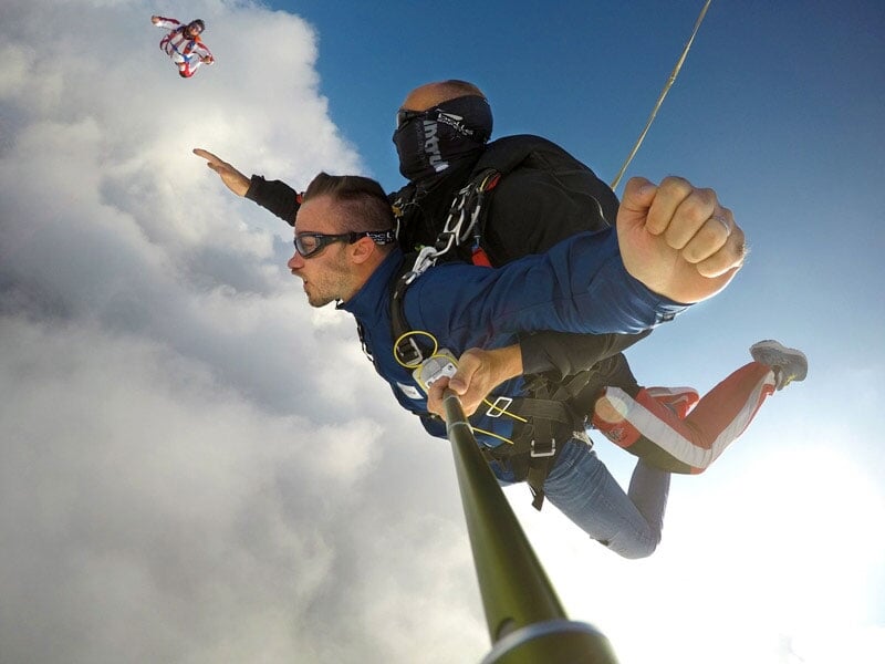 Deux hommes en parachute en tandem sautent d'un avion, l'un tenant un perche selfie, ciel bleu et nuages en arrière-plan.