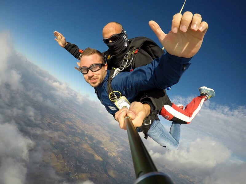 Deux hommes en parachute en tandem sautent d'un avion au-dessus de la campagne avec ciel bleu et nuages en arrière-plan.