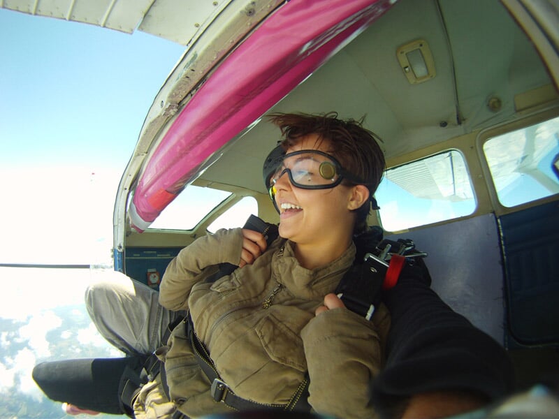 Une personne souriante portant des lunettes de protection est assise à l'intérieur d'un avion avant de sauter en parachute, prête pour l'aventure.