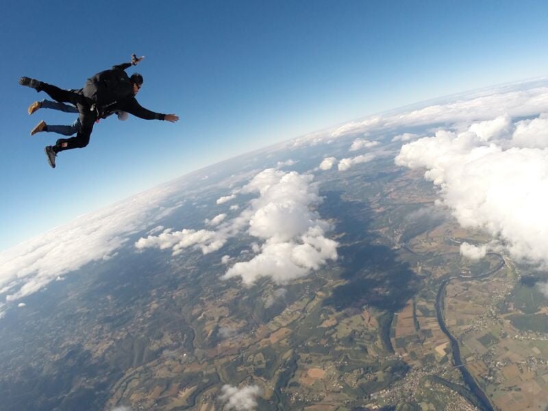 Deux personnes en chute libre lors d'un saut en parachute au-dessus d'un paysage rural avec des nuages et un ciel bleu clair.