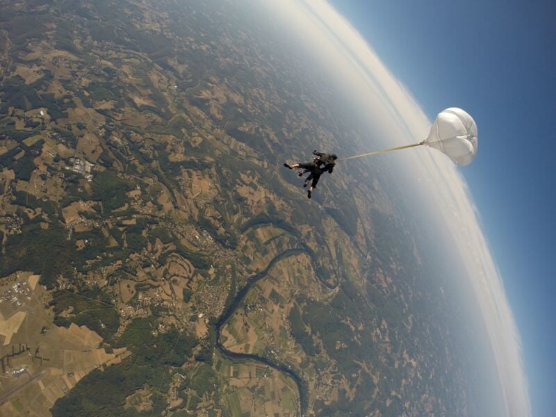 Deux personnes en parachute en tandem survolant un paysage vallonné avec rivières et champs sous un ciel clair.
