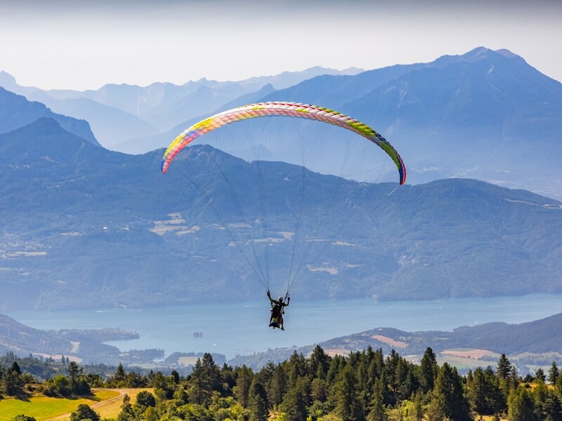 Parapente en tandem en plein vol au-dessus d’un paysage montagneux avec lac, forêts et collines, sous une voile colorée en arc. Vue panoramique sur les reliefs en arrière-plan.