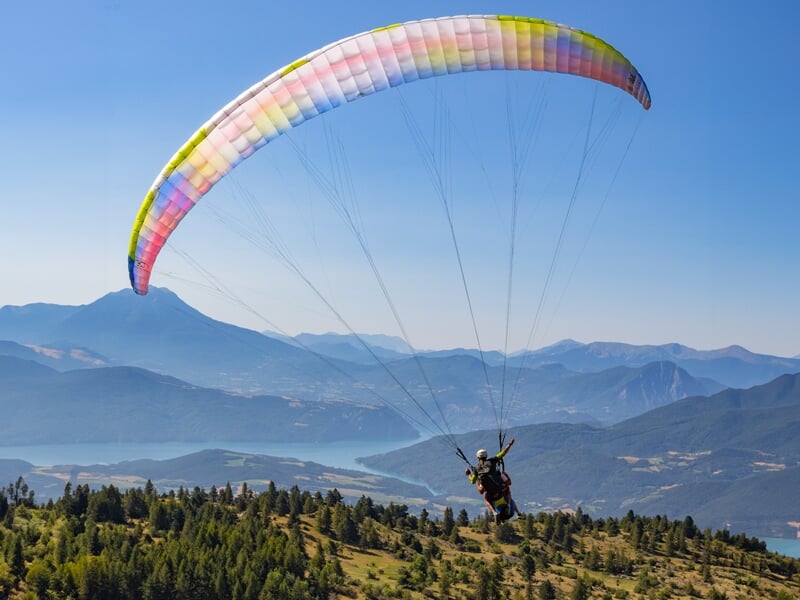 Vol en parapente biplace au-dessus d’une crête boisée avec vue dégagée sur les montagnes et un grand lac en contrebas. La voile multicolore est bien gonflée dans un ciel limpide.