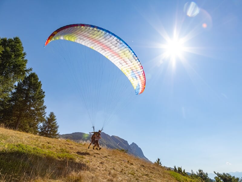 Décollage en tandem d’un parapente sur une pente ensoleillée, voile arc-en-ciel déployée au-dessus des deux personnes courant vers le vide. Soleil éclatant dans un ciel bleu dégagé.