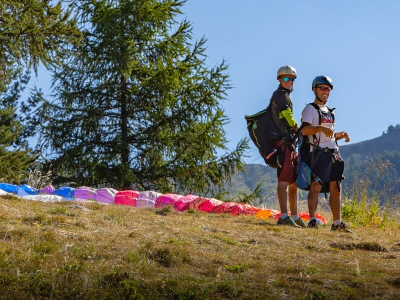 Deux parapentistes en tenue de vol et casque se tiennent sur une pente herbeuse, prêts pour un décollage. La voile multicolore est déployée au sol derrière eux, avec des sapins en arrière-plan.