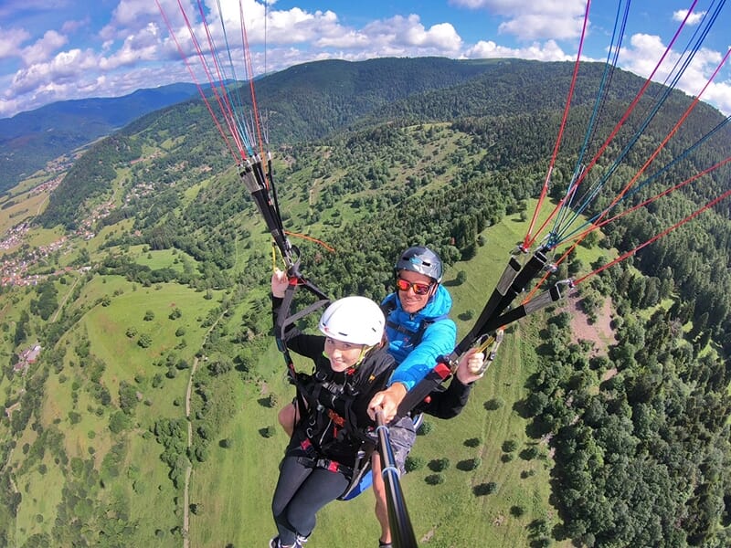 Un vol en tandem en parapente au-dessus de collines verdoyantes, les deux pilotes souriants regardent la caméra, suspendus sous leur voile, avec un panorama de montagnes et de vallées verdoyantes en a