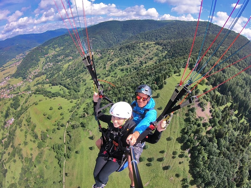 Deux personnes en parapente en tandem survolent une vallée verdoyante sous un ciel bleu avec quelques nuages blancs.