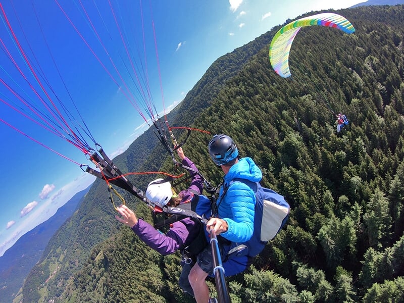 Un vol en parapente en tandem capturé en pleine action, les deux pilotes survolent une dense forêt de conifères. Une autre voile multicolore est visible en arrière-plan, planant au-dessus des arbres e