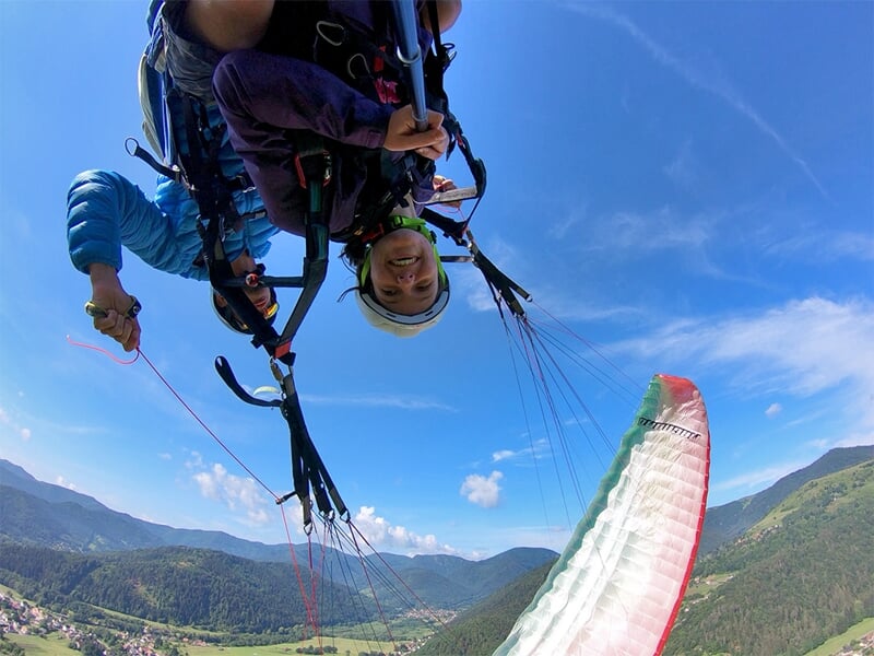 Une personne souriante suspendue dans un harnais, capturée en plein vol lors d'un vol en parapente acrobatique, avec la voile en arrière-plan sous un ciel bleu.