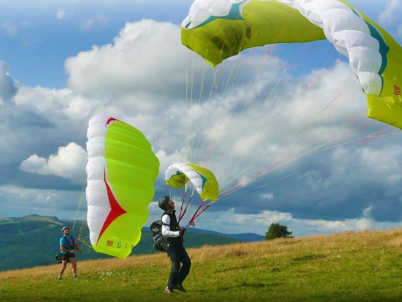 Deux parapentistes manœuvrent leurs voiles vertes et blanches sur une colline herbeuse, avec des nuages et des montagnes à l'arrière-plan, dans une scène de préparation avant le décollage.