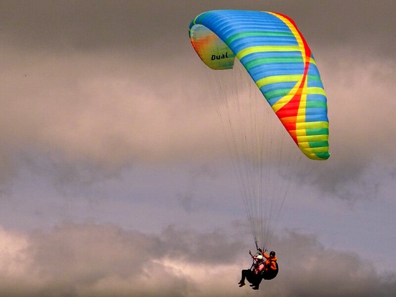 Un parapente biplace aux motifs multicolores (dominantes de bleu et jaune) plane dans les airs sous un ciel nuageux, les deux pilotes sont suspendus dans le harnais.