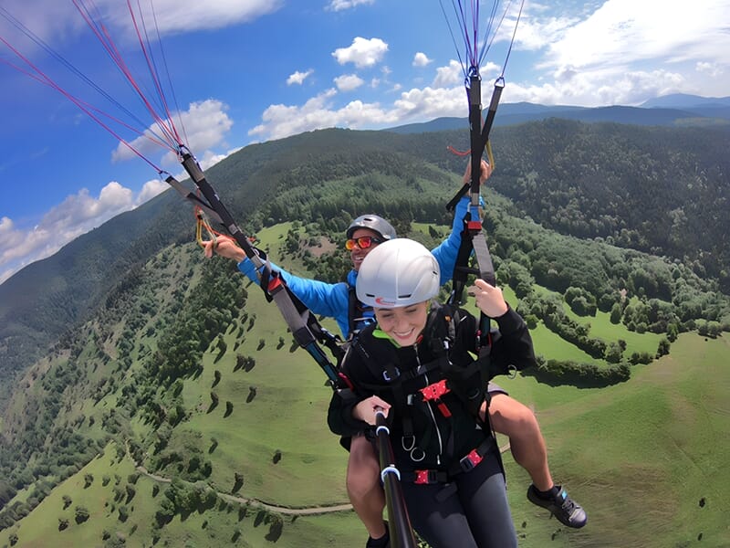 Personne en tandem avec un instructeur lors d'un baptême de parapente au-dessus d'une vallée verdoyante sous un ciel bleu